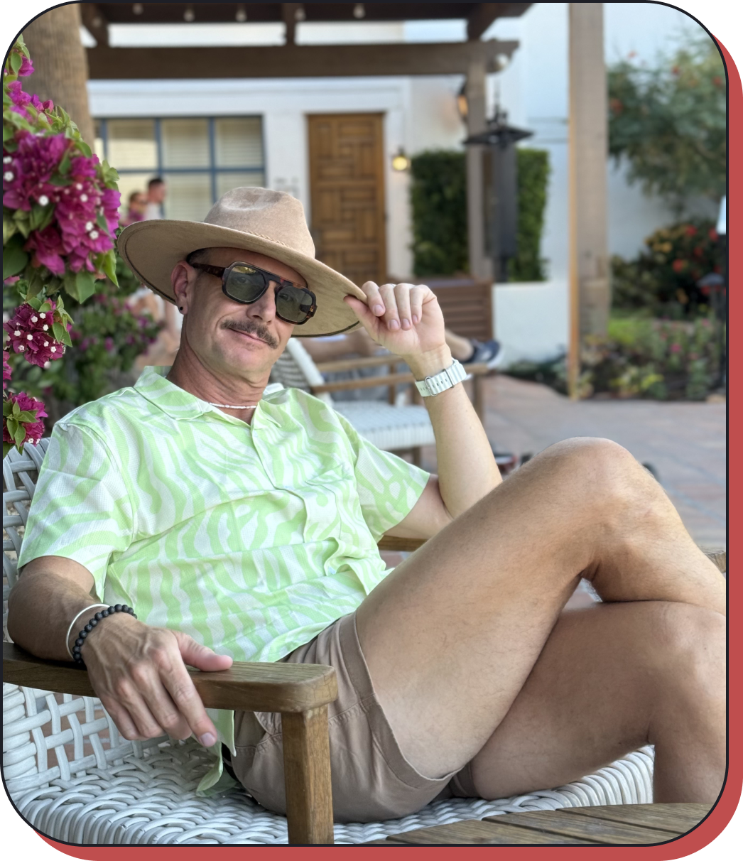 A man relaxing outdoors on a patio, wearing a wide-brimmed hat, sunglasses, a green patterned shirt, and beige shorts, sitting on a wicker chair near pink flowers and greenery, with a building and other people in the background.
