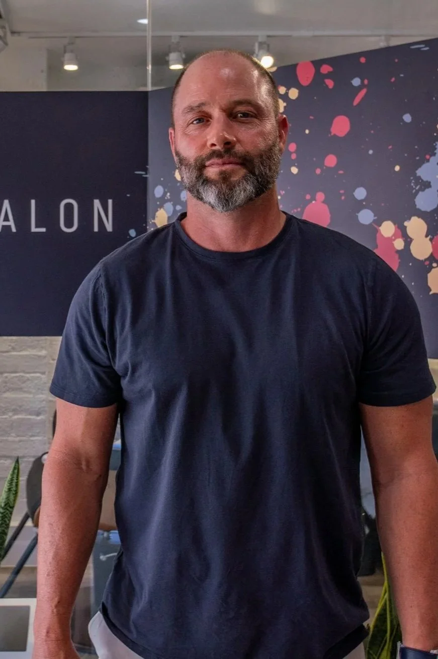 A middle-aged man with a beard and mustache, wearing a dark blue t-shirt, standing indoors with a colorful abstract background and a partial sign that reads 'ALON'.