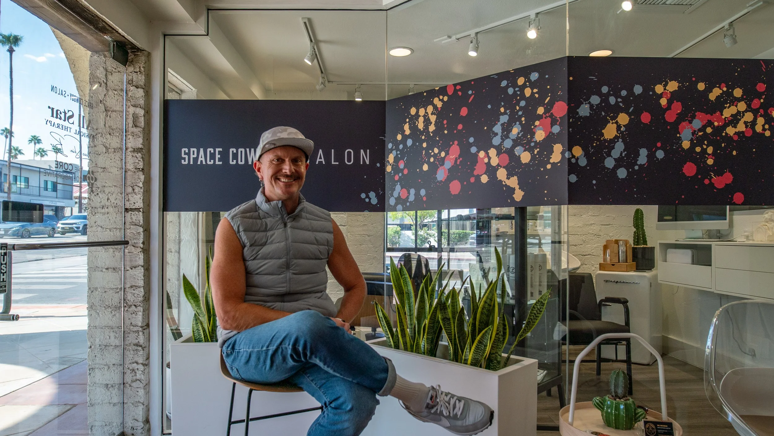A man sitting on a chair inside a modern space office with plants and windows, smiling at the camera. There is a sign that reads "Space Coworking" in the background.