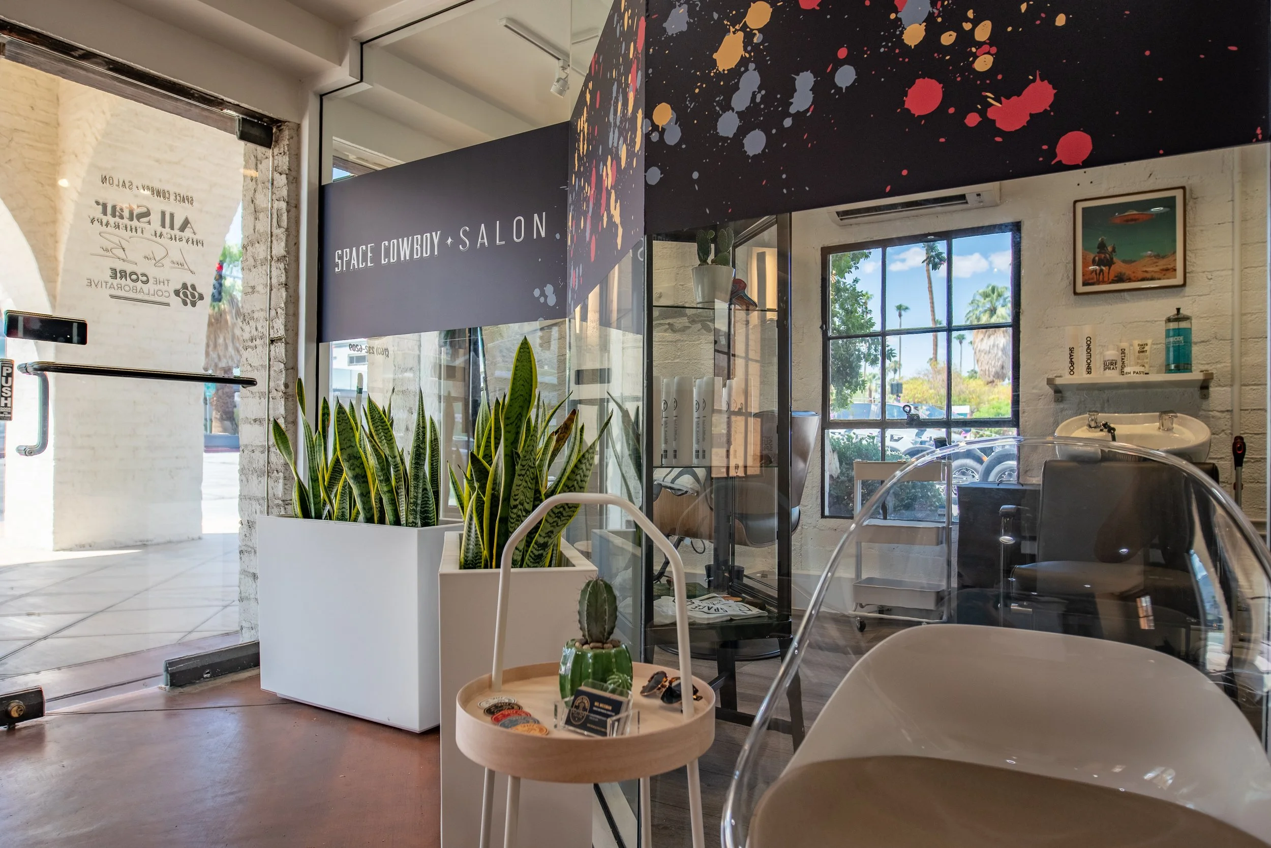 Interior of a salon with a sign that reads 'SPACE COWBOY SALON,' a large planter with tall green plants, a small round table with decorative items, and a sink area with a window showing palm trees outside.
