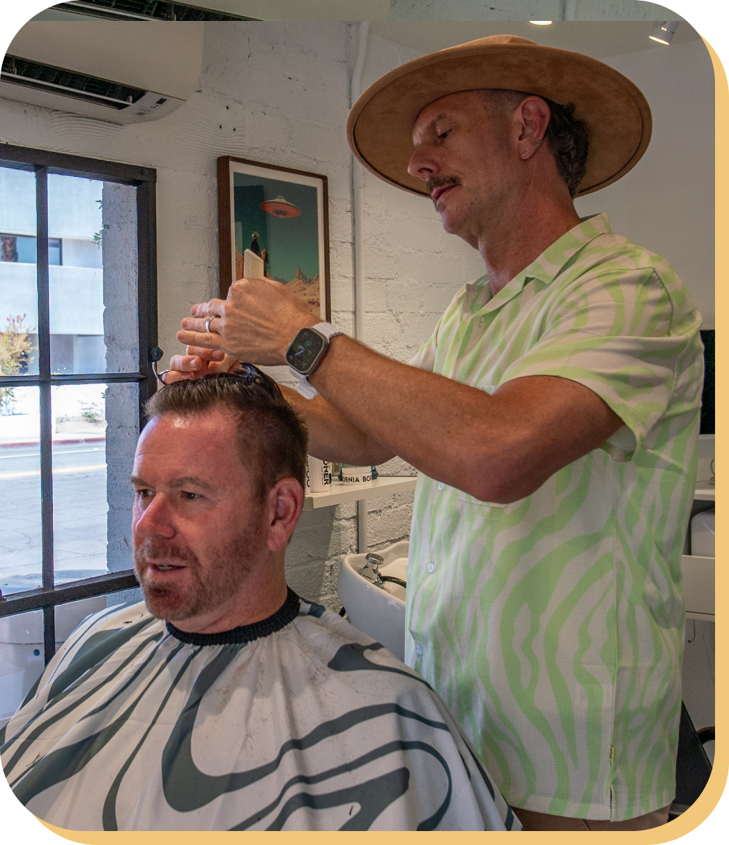 A man receiving a haircut from a barber in a modern salon with white brick walls and a window. The barber, wearing a wide-brimmed hat and a light-colored, patterned shirt, is using scissors to cut the client's hair. The client is seated in a barber chair, covered with a cape, and has short, dark hair.